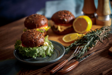 Dark burger with grain bread on dark ceramic plate, salad, rosemary and lemon