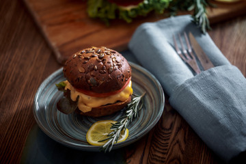 Dark burger with grain bread on dark ceramic plate, salad, rosemary and lemon