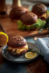 Dark burger with grain bread on dark ceramic plate, salad, rosemary and lemon