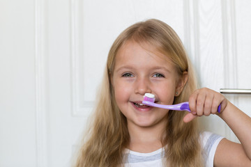 Portrait of cute little girl with blonde hair which cleaning tooth with brush and toothpaste in bathroom 