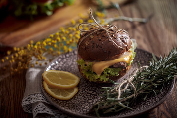 Dark burger with grain bread on dark ceramic plate, salad, rosemary and lemon