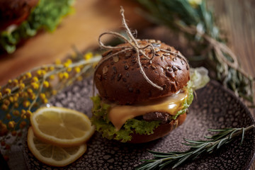 Dark burger with grain bread on dark ceramic plate, salad, rosemary and lemon