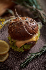 Dark burger with grain bread on dark ceramic plate, salad, rosemary and lemon