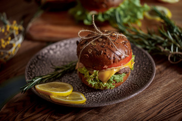 Dark burger with grain bread on dark ceramic plate, salad, rosemary and lemon