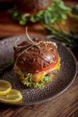 Dark burger with grain bread on dark ceramic plate, salad, rosemary and lemon