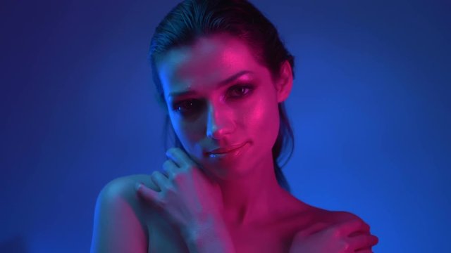 Glowing fashionable model with glitter makeup in blue and pink neon lights cuddling herself and watching into camera with calm smile.