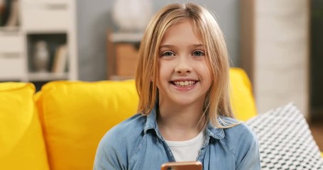 Portrait of the joyful Caucasian beautiful teenage small girl scrolling and texting sms on the smartphone and smiling to the camera. Close up. At home.