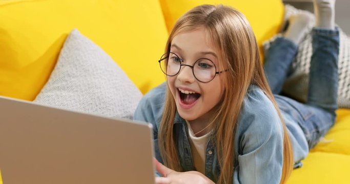 Pretty and joyful Caucasian cute blond girl in glasses watching something on the laptop computer and doing surprised and happy face while lying on the sofa. Indoor. Close up.