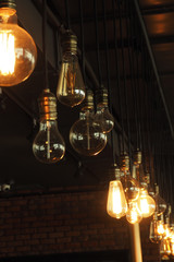 incandescent bulbs of different shapes and sizes hanging from a black ceiling and a brick wall...