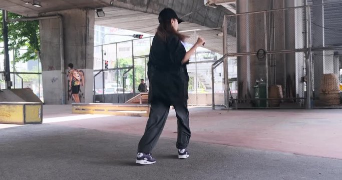 Wide angle of young woman doing freestyle street dancing with skateboard park and unidentified people in background. Asian woman.