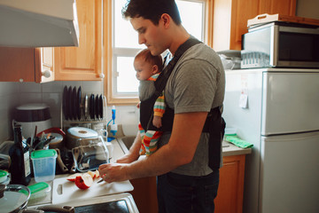 Young Caucasian father with newborn baby in carrier preparing lunch. Man parent carrying child...