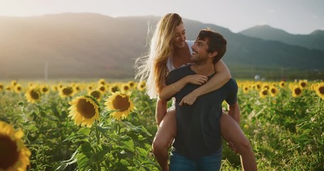 Happy young attractive couple in sunflower field together at sunset in slow motion, happy attractive woman riding on her boyfriend's back, summer romance lifestyle