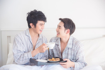 Young Asian Gay couple wearing pajamas sitting on the bed and having breakfast together ,Homosexual couple concept.