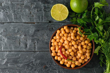 Clay bowl filled with boiled chickpeas on a black wooden table with parsley and lime. The view from the top.