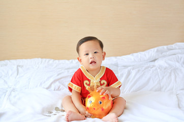 little Asian baby boy in traditional Chinese dress with a piggy bank sitting on bed at home. Kid saving money concept.
