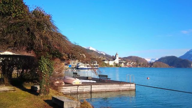 Walk by the Wolfgangsee, enjoy the lakeside park, watch the bright blue rippled surface of the lake, ferries in port and the Pilgrimage church on background, St Wolfgang, Salzkammergut, Austria