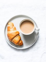 Simple breakfast or snack - coffee with milk and a classic croissant on a light background, top view