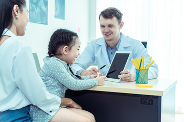 Fototapeta premium Young friendly caucasian pediatrician doctor is examining child patient girl with her mother ,consultation with a stethoscope and tablet pc in clinic office .Healthcare and medical kid concept