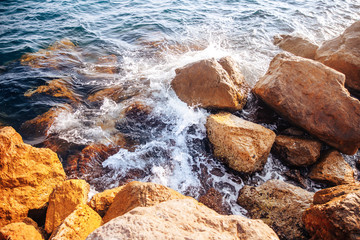 Stormy sea with waves and splashes breaks on sharp stones, warm day, light of sun