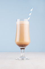 Cappuccino coffee cocktail with cream and striped straw in transparent glass on pastel blue wall and white wood board, vertical.