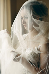 Beautiful young bride holding veil in white wedding dress, portrait of brunette bride posing near window in hotel room, morning before the wedding