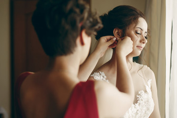 Beautiful bridesmaid helping brunette bride in white wedding dress put on luxury earrings, morning...