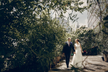 Romantic newlywed couple walking in the park, handsome groom and happy beautiful bride with bouquet holding hands in sunny botanic garden