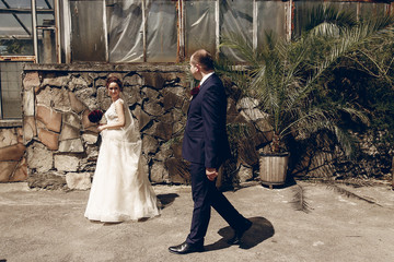 Happy handsome groom walking with beautiful brunette bride in white wedding dress with bouquet near stone wall in botanic garden