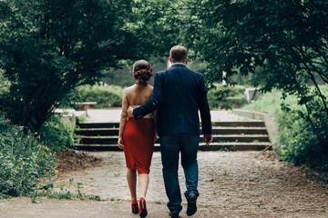 Obraz premium Romantic newlywed couple walking in park, handsome goom holding hand around waist of sexy bride in red skirt, romance concept