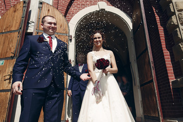 Happy smiling bride and groom walking out of church after wedding ceremony, newlyweds throwing rice at stairs near cathedral moment, marriage concept