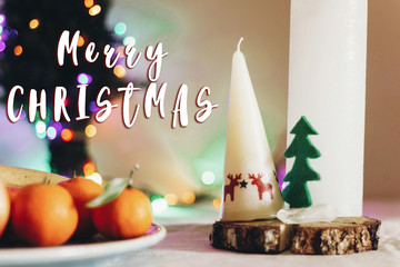 merry christmas text sign on christmas rustic table with candle with reindeers and felt tree and fruits on colorful lights background. space for text. seasonal greetings holidays card concept