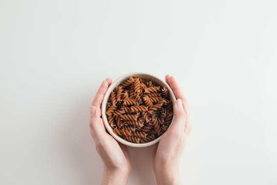 Hands Holding Bowl Of Whole Grain Fusilli Pasta, White Background, Free Space For Text, Horizontal