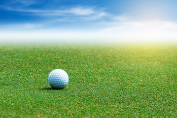 White Golf ball on the green grass on blurred blue sky with clouds background.