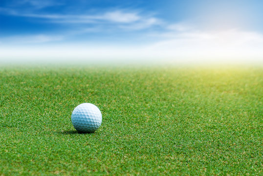 White Golf ball on the green grass on blurred blue sky with clouds background.
