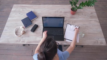 overhead view of freelancer girl typing text on laptop keyboard and writes notes in clipboard sitting at the table during studying online