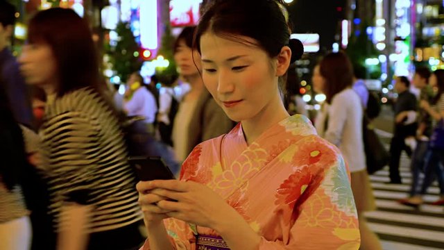 Attractive Japanese woman in a kimono messing on a cell phone. Traffic and the lights of Shinjuku are behind her.