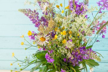 Fototapeta na wymiar Bouquet of wild and meadow flowers from lupines, buttercups, a camomile and a fresh grass against the background of a wooden background of blue color