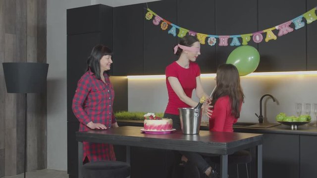 Happy woman celebrating Birthday with mother and best friend in domestic festively decorated kitchen. Surprised female with blindfold groping friend coming to congratulate her with anniversary at home