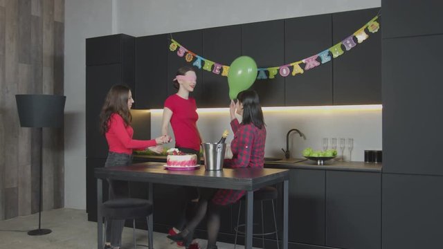 Cheerful adult female enjoying celebration of Birthday in narrow circle with mother and good friend in festively decorated kitchen. Prepared for surprise blindfolded female groping and hugging mom.