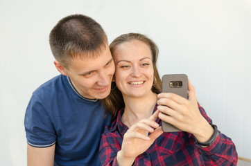 guy with short haircut in blue T-shirt and s,iling girl with dark long hair in red and blue checkered shirt take selfie on the phone