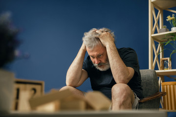 terrible ordeals. Old bearded man with alzheimer desease sitting on the chair and suffering from headache. Concept of illness, memory loss due to dementia, healthcare, depression, stress out.