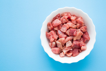 Red, raw, fresh meat pieces in white bowl on light pastel blue table. Closeup. Empty place for text. 