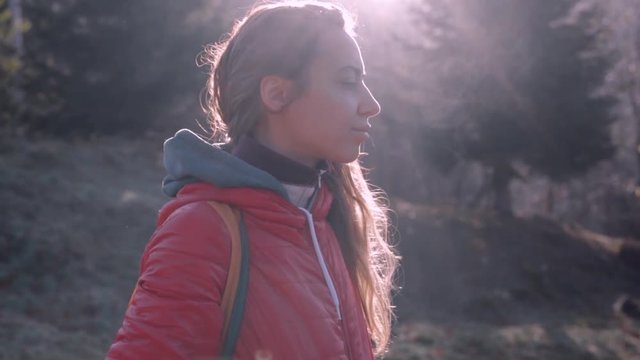 pretty woman hiker in red jacket with small orange backpack is standing in a woods at early cold morning, breathes deeply and exhales steam, wonderful backlight from sun on background. admired woman
