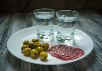 Fototapeta na wymiar Two shots of vodka, olives and salami slices on white plate