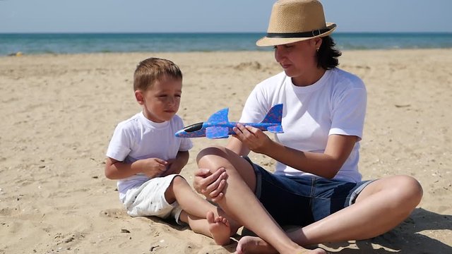 A little cute boy and his mom are sitting on the sandy seashore on a sunny summer day. Mom holds a toy plane and plays with the child.