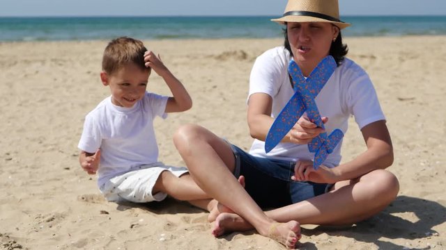 A happy mother and her son are sitting on a sandy beach on a sunny summer day and playing with a toy plane, in slow motion