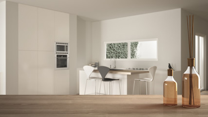 Wooden table top or shelf with aromatic sticks bottles over blurred modern white kitchen with parquet floor and island with stools, white architecture interior design