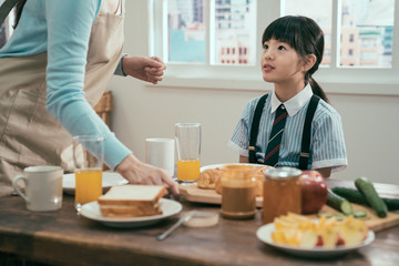unrecognized mom in apron standing at wooden table with healthy meal. cute little girl in uniform looking at mother waiting for delicious breakfast in morning before school. sweet family time.