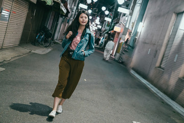 Full length portrait of stylish hipster girl backpacker looking aside while standing in urban city osaka japan. woman going home relaxing on street with no traffic quiet illumination lights at night