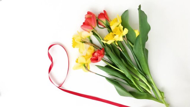 Red ribbon in the shape of a heart is unwound. White desktop with spring flowers flat lay, yellow daffodils and red tulips. Bouquet for St Valentine's day, Mother's day or birthday top view